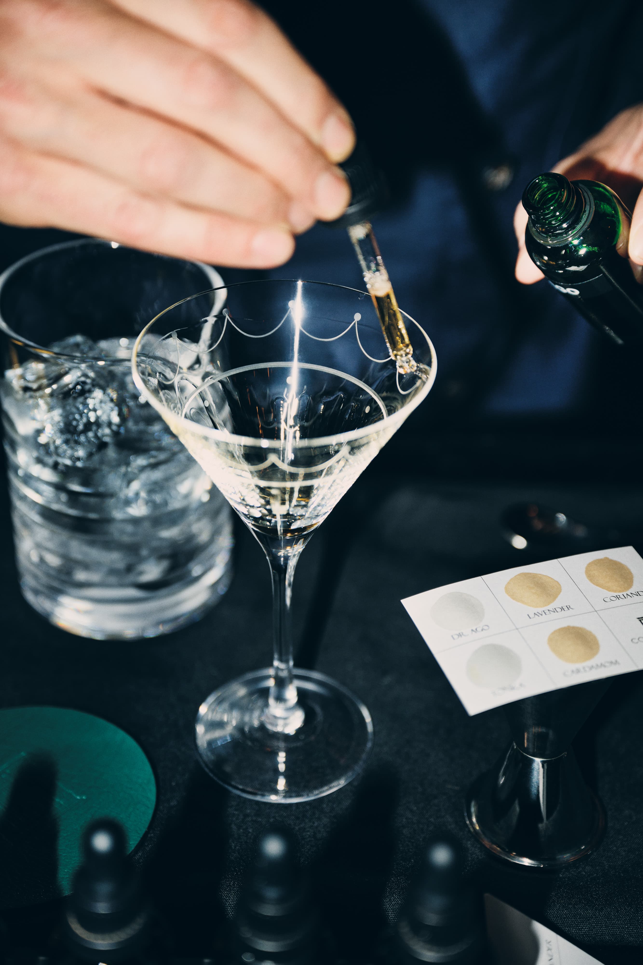 An empty martini glass with aromatics being added from a dropper.