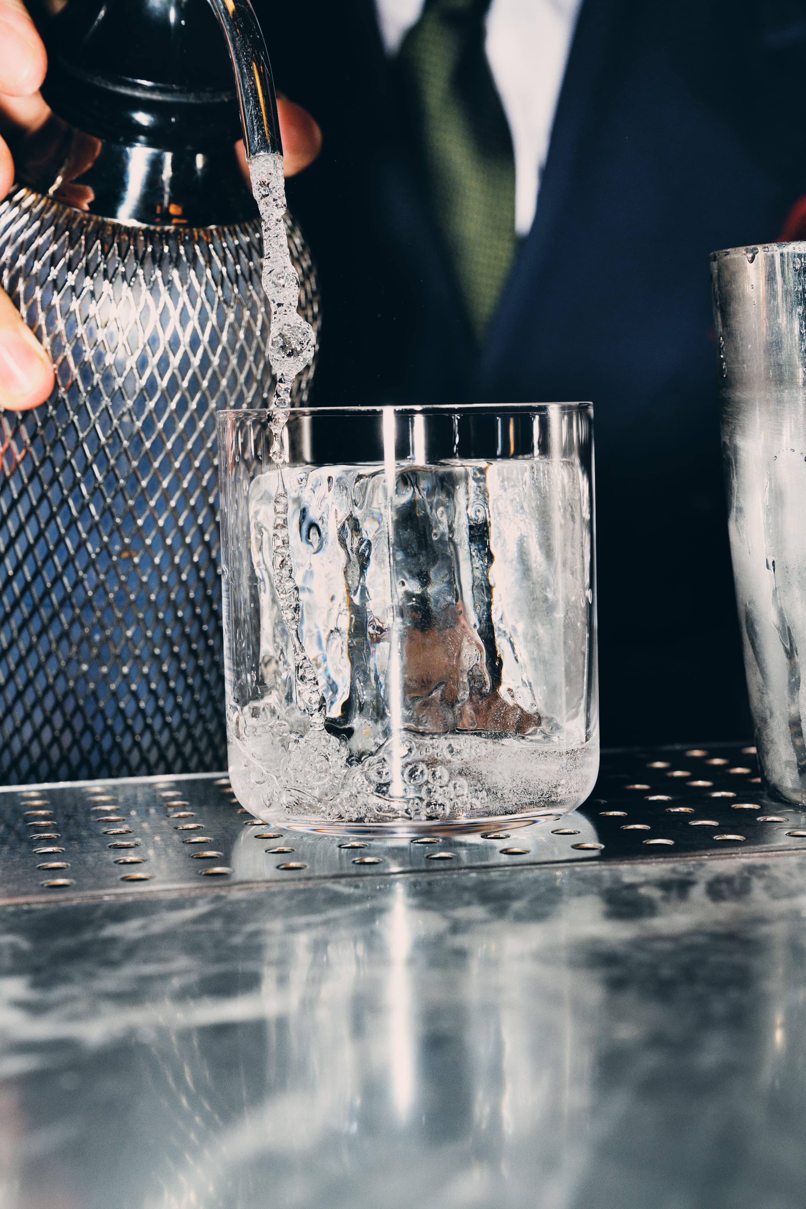 A person pouring a cocktail from a shaker into a glass with one large ice cube