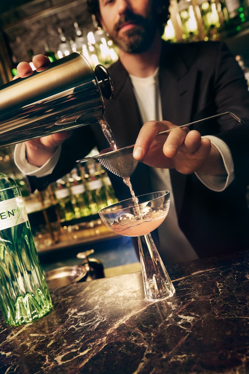 Matteo Di ienno pouring a cocktail shaker through a strainer into a coupette glass.