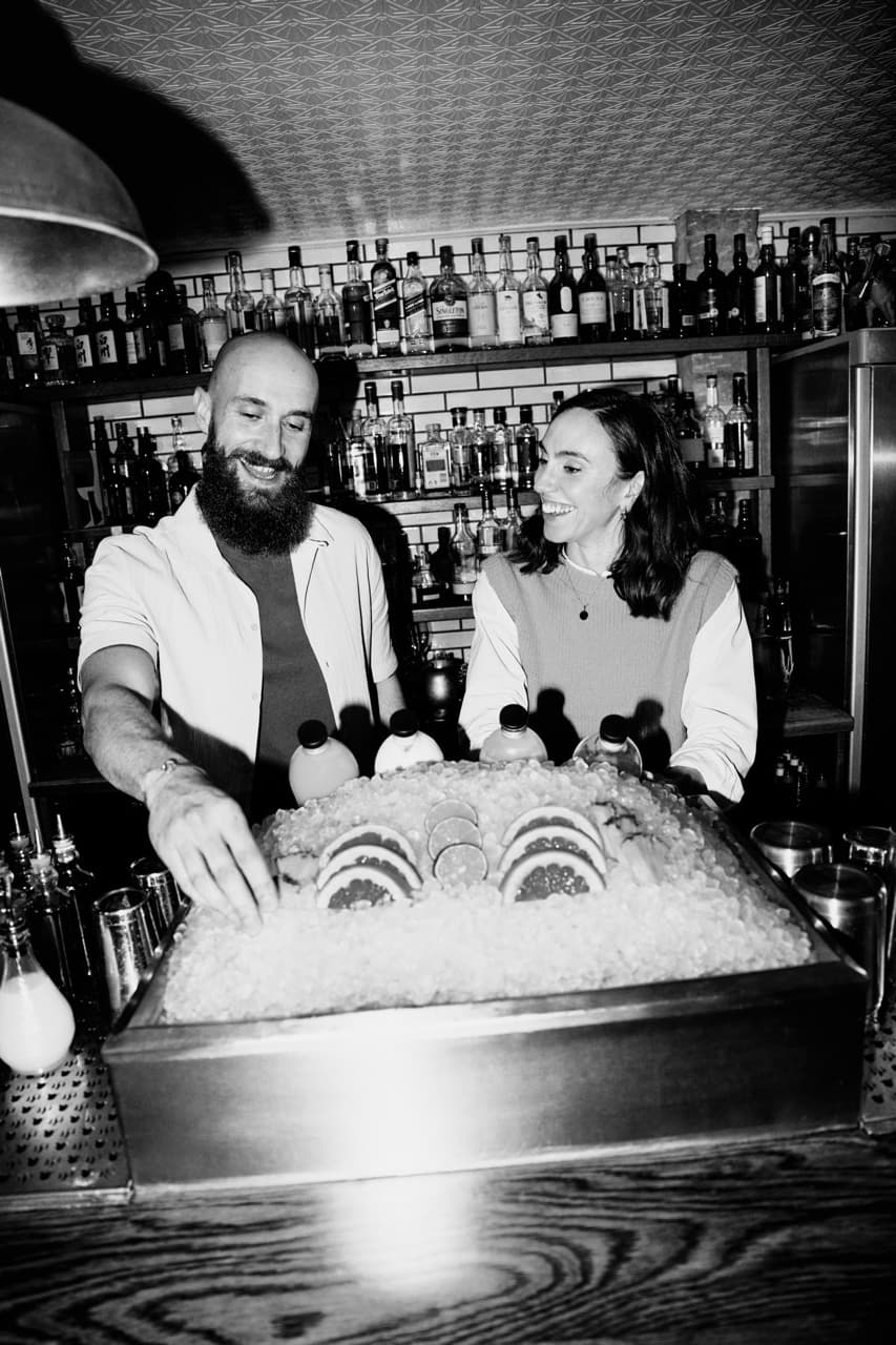 Keila & Yoann standing behind an ice box of garnishes, including grapefruit