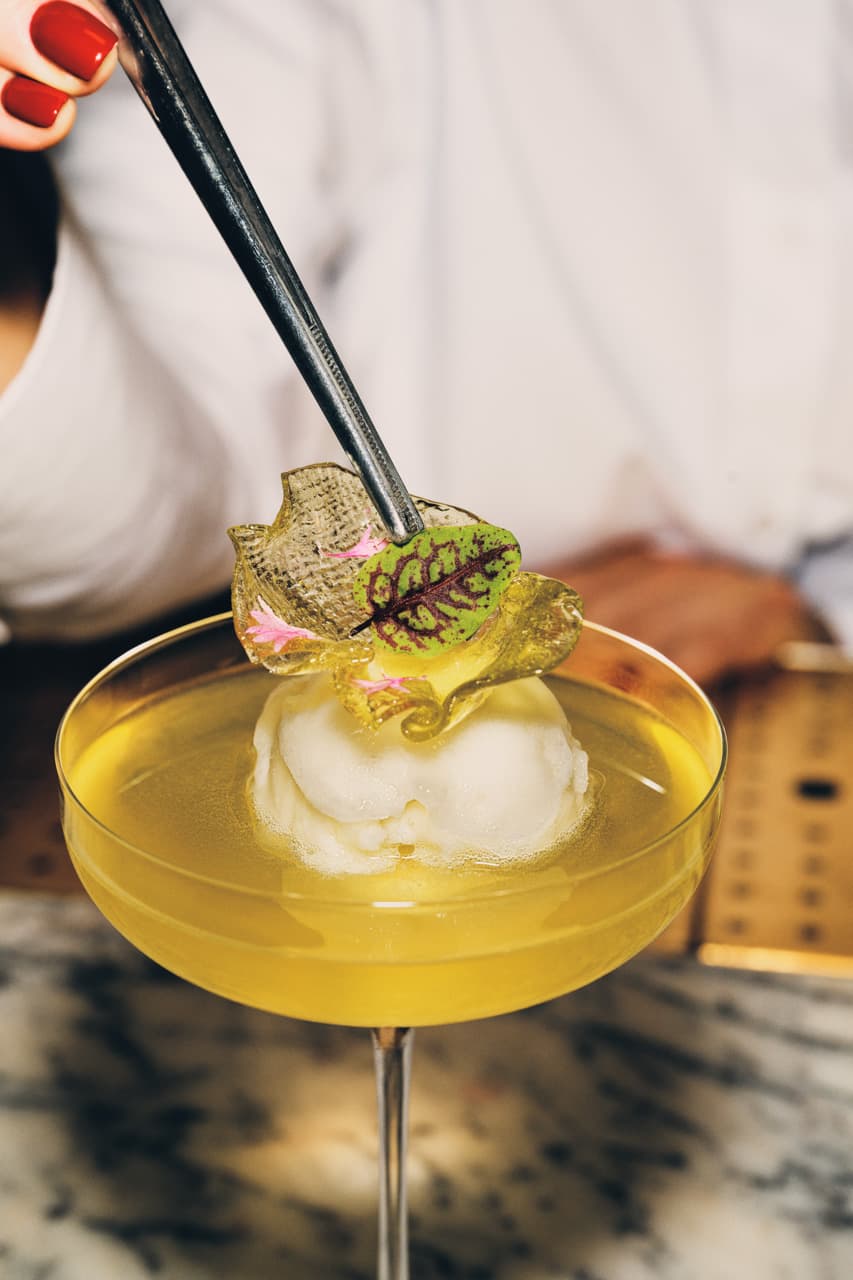 A cocktail glass sits on a marble counter, close behind we see the arm and shoulders person holding some tweezers to place the final flourish of a small green leaf onto the cocktail.