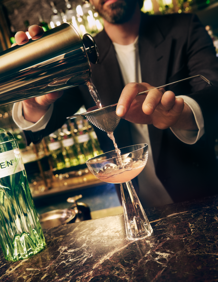 A hand pouring a drink from a cocktail shaker through a strainer, in to a cocktail glass.